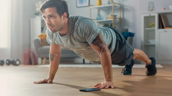 Athletic Fit Man póló és rövidnadrág csinál push up gyakorlatok használata közben stopper az ő Phone. Ő képzés otthon az ő Living Room minimalista Interior. — Stock Fotó