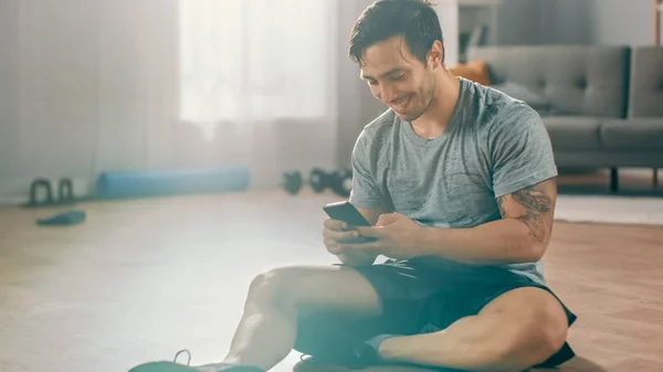 Gelukkig sterke atletische fit man in T-shirt en shorts is met behulp van een mobiele telefoon na Ochtendoefeningen thuis in zijn ruime en lichte woonkamer met minimalistisch interieur. — Stockfoto