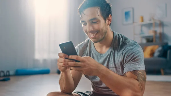 Happy Strong Athletic Fit Man in T-shirt e pantaloncini sta utilizzando un telefono cellulare dopo gli esercizi mattutini a casa nel suo ampio e luminoso soggiorno con interni minimalisti . — Foto Stock