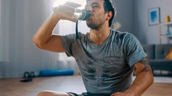 Gelukkige en tevreden sterke atletische fit man in T-shirt en shorts is het drinken van water na Ochtendoefeningen thuis in zijn ruime en lichte woonkamer met modern interieur. — Stockfoto