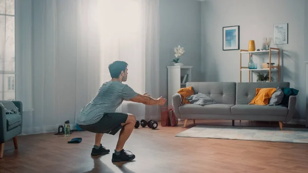 Строгий атлетичный мужчина в футболке и шортах выполняет упражнения на корточках дома в своем просторном и ярком костюме с интерьером. . — стоковое фото