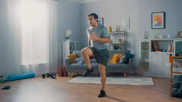 Strong Athletic Fit Man w T-shirt i szorty jest energicznie jogging w miejscu w domu w jego przestronnym i jasnym salonie z minimalistycznym wnętrzem. — Zdjęcie stockowe