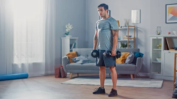 Gespierde atletische fit man in T-shirt en shorts doet oefeningen met dumbbells thuis in zijn ruime en lichte woonkamer met minimalistisch interieur. — Stockfoto