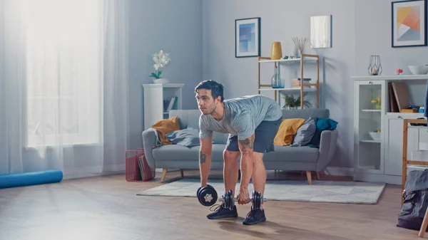 Muscular Atlético Fit Man em T-shirt e Shorts está fazendo exercícios com halteres em casa em sua espaçosa e brilhante sala de estar com interior minimalista . — Fotografia de Stock