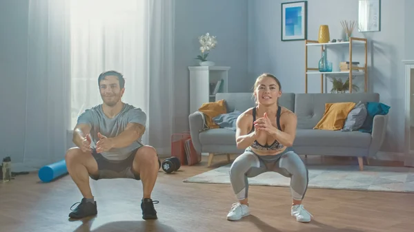 Homem atlético muscular e bela mulher fitness em roupas de treino estão fazendo exercícios de agachamento em seu apartamento brilhante e espaçoso com interior minimalista . — Fotografia de Stock