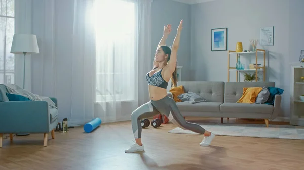 Hermosa chica de fitness segura en una parte superior atlética está haciendo ejercicios de yoga de estiramiento en su brillante y espaciosa sala de estar con interior minimalista . —  Fotos de Stock