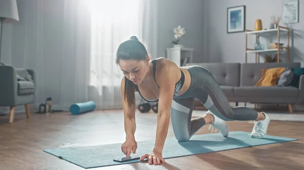 Chica fuerte y segura de fitness en ropa deportiva atlética gris está haciendo ejercicios de entrenamiento push up mientras usa un cronómetro en su teléfono. Ella está entrenando en casa en su brillante sala de estar con interior acogedor . —  Fotos de Stock