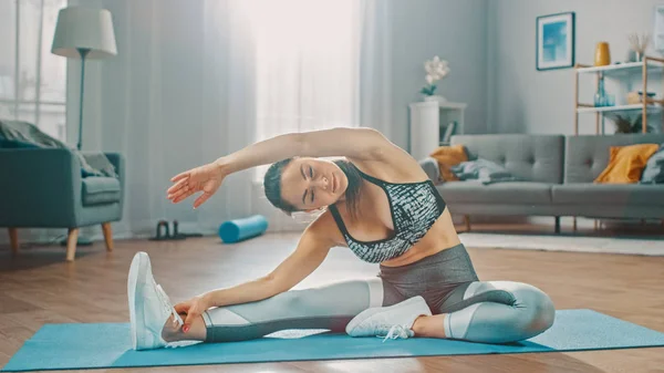 Hermosa chica de fitness tetona en una parte superior atlética está haciendo ejercicios de yoga de estiramiento en su brillante y espaciosa sala de estar con interior moderno . — Foto de Stock