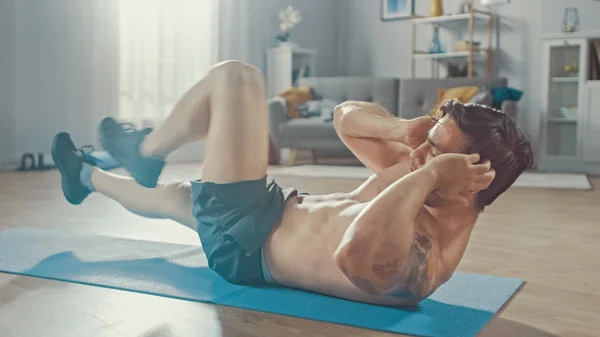 Sterke atletische shirtless fit man in grijze shorts doet buik oefeningen thuis in zijn ruime en zonnige woonkamer met minimalistisch interieur. — Stockfoto