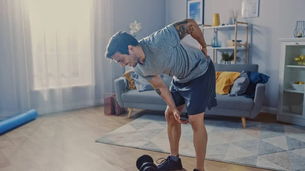 Muscular atlético Shirtless Fit Man em calções cinza fere as costas depois de levantar halteres em casa em sua espaçosa e ensolarada sala de estar com interior minimalista . — Fotografia de Stock