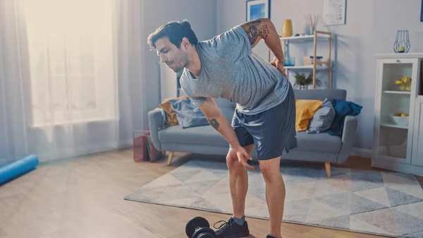 Muscular atlético Shirtless Fit Man em calções cinza fere as costas depois de levantar halteres em casa em sua espaçosa e ensolarada sala de estar com interior minimalista . — Fotografia de Stock
