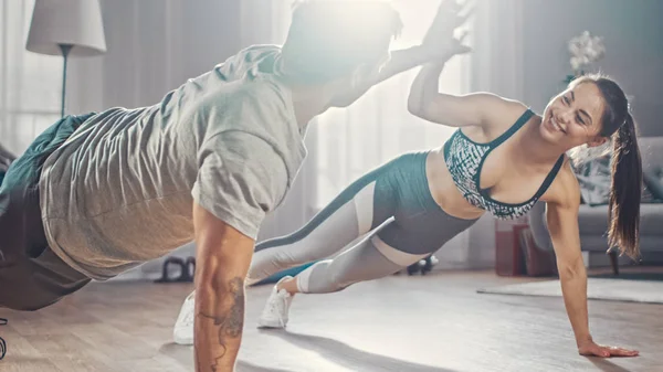 Smiling Strong and Beautiful Athletic Fitness Couple in Workout Clothes Doing Push Up Exercises and Giving Each Other a High Five in Their Bright and Spacious Living Room with Minimalistic Interior.