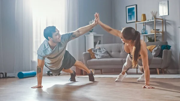 Ισχυρή και όμορφη αθλητική Fitness ζευγάρι στην προπόνηση ρούχα κάνει push up ασκήσεις και δίνοντας ο ένας στον άλλο ένα ψηλό πέντε στο φωτεινό και ευρύχωρο σαλόνι τους με μινιμαλιστικό εσωτερικό. — Φωτογραφία Αρχείου