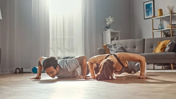 Shot van een sterke en mooie atletische fitness paar in workout kleren doen push up oefeningen. Lichte en ruime woonkamer met minimalistisch interieur. — Stockfoto