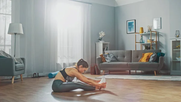 Hermosa chica de fitness segura en una ropa de entrenamiento atlético haciendo ejercicios de estiramiento en su brillante y espaciosa sala de estar con interior minimalista acogedor . — Foto de Stock