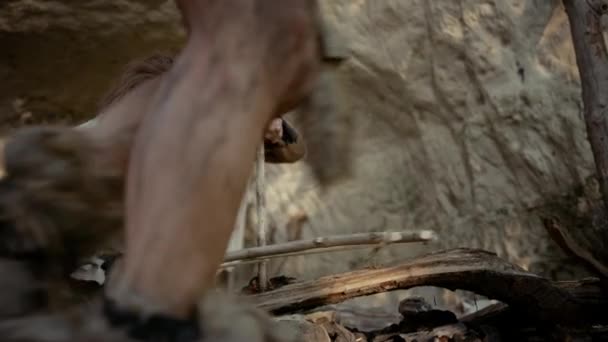Первобытный пещерный человек в шкуре животного пытается разжечь огонь с помощью метода лука. Неандерталец Киндл Первый рукотворный пожар в истории человеческой цивилизации. Разведение огня для приготовления пищи. Slow Motion Arc Shot — стоковое видео