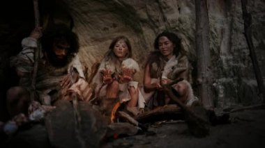 Tarih Öncesi İlkel Avcı-Toplayıcılar kabilesi Hayvan Derileri Giyen Bir Mağarada Gece Yaşıyor. Neanderthal ya da Homo Sapiens Ailesi Şenlik Ateşi 'nde Isınmaya Çalışıyor, Ateşin Üzerinde Elele tutuşuyor, Yemek Pişiriyor