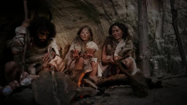 動物の皮を身に着けている先史時代の原始の狩猟採集民の部族は、夜に洞窟に住んでいます。ネアンデルタール人やホモ・サピエンスの家族はたき火で暖かくしようとしています,火の上に手を保持,料理の食べ物 — ストック動画