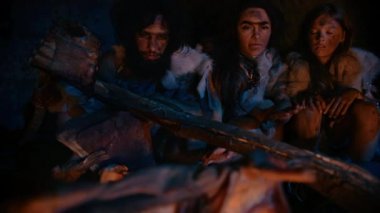 Neandertal veya Homo Sapiens Aile Pişirme Hayvan Eti Bonfire üzerinde ve sonra yeme. Prehistorik Avcı-Toplayıcılar Kabilesi Gece Karanlık Korkunç Mağarada Yemek Hayvan Derileri Giyen. Yakın Çekim