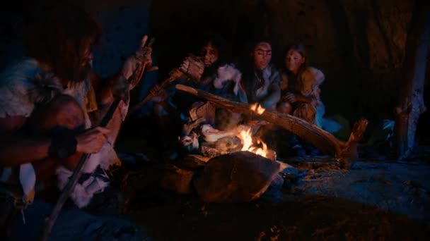 Neandertaler oder Homo sapiens Familie kocht Tierfleisch über dem Lagerfeuer und isst es dann. Stamm prähistorischer Jäger und Sammler in Tierhäuten, die nachts in einer dunklen Gruselhöhle essen. Zoom in Schuss