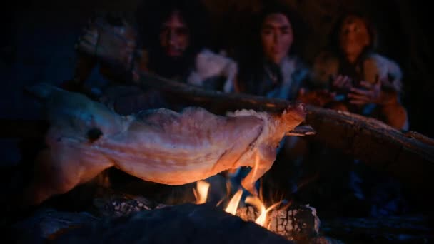 Neanderthal o Homo Sapiens Famiglia cucinare carne animale sopra il falò e poi mangiarlo. Tribù di cacciatori-raccoglitori preistorici che indossano pelli di animali che mangiano in una grotta oscura e spaventosa di notte. Primo piano girato — Video Stock