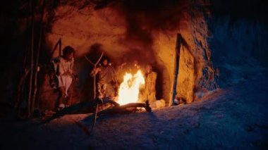 Tarih öncesi avcı-toplayıcı kabilesi hayvan derileri giyip geceleri mağaranın dışında şenlik ateşi etrafında dikiliyor. Neandertal / Homo Sapiens Ailesi Pagan Dini Ayini Yakındaki Ateşin Portresi