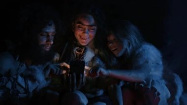 Tarih Öncesi, İlkel Avcı-Toplayıcılar kabilesi Hayvan Derisi Giyen Bir Mağarada Gece Akıllı Telefon Kullanıyor. Neanderthal / Homo Sapiens Ailesi Cep Telefonu 'nda İnternet Taraması, Ateşin Üzerinde Yemek Pişirme