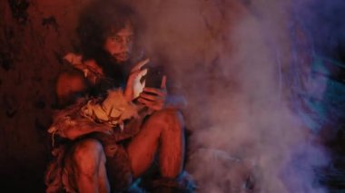 Tarih öncesi, ilkel avcı toplayıcı hayvan derisi giyen kabile geceleri mağarada akıllı telefon kullanır. Cep telefonunda Neandertal / Homo Sapiens Erkek Tarayıcı İnternet, Videolar İzliyor