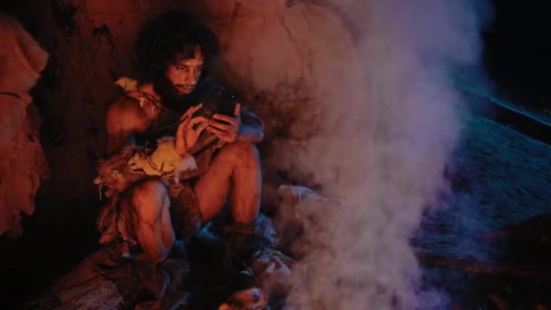 Tribe of Prehistoric, Priforcious Hunter Gatherer Wearing Animal Skin Skin Uses Smartphone in a Cave at night. 네안데르탈인 / 호모 사피엔 인 Male Browsing Internet on Mobile Phone, Watches Videos — 비디오