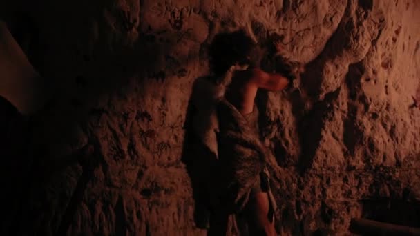 動物の皮を身に着けている原始的な先史時代のネアンデルタールは、夜の壁に動物と抽象を描きます。ペトログリフ、火によって照らされた岩の絵画で最初の洞窟アートを作成します。バックビューズームイン — ストック動画