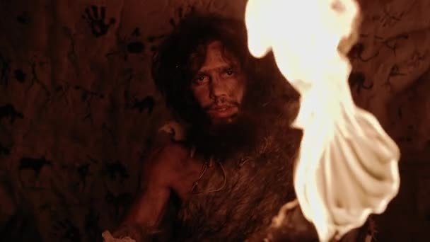 Портрет первозданної печери Caveman, що носить шкіру тварин, стоїть у своїй печері вночі, тримає факел з вогнем. Примітивний неандертальський мисливець / Homo Sapiens At Night Alone. На задньому плані печерні малюнки — стокове відео