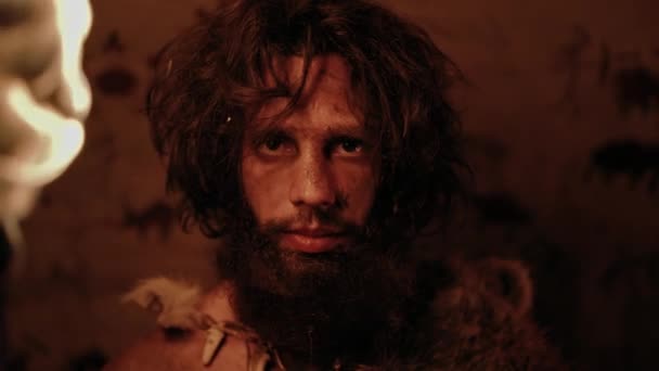 Портрет первозданної печери Caveman, що носить шкіру тварин, стоїть у своїй печері вночі, тримає факел з вогнем. Примітивний неандертальський мисливець / Homo Sapiens At Night Alone. На задньому плані печерні малюнки — стокове відео