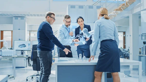 Diverse Team of Industrial Robotics Engineers Gathered Around Table With Robot Arm, They Use Tablet Computer to Manipulate and Program it to Pick Up and Move Metal Component. Bright Facility