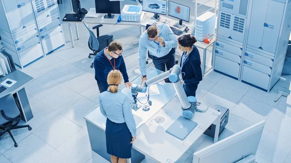 Team of Industrial Robotics Engineers Gathered Around Table With Robot Arm, They Use Tablet Computer to Manipulate and Program it to Pick Up and Move Metal Component. Top Down Above Shot