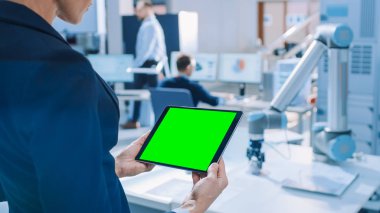 Mühendis Yeşil Mock-up Ekran Dijital Tablet Bilgisayar tutar. Modern Parlak Araştırma Tesisinde Fonksiyonel Robot Kolu ile Çalışan Endüstri Mühendislerinden Oluşan Arka Plan Ekibinde