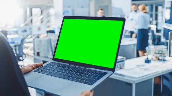 Μηχανικός κρατά πράσινη μακέτα υπολογιστή-επάνω οθόνη laptop. Στο παρασκήνιο της ομάδας βιομηχανικών μηχανικών που εργάζονται με λειτουργικό χέρι ρομπότ στο σύγχρονο φωτεινό ερευνητικό κέντρο — Φωτογραφία Αρχείου