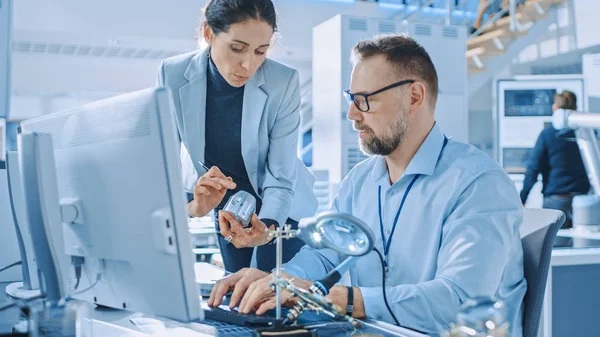 Ingeniero industrial femenino habla con el especialista en electrónica, sostiene el prototipo del componente del metal, él trabaja en computadora. Oficina moderna y brillante con gente con estilo — Foto de Stock