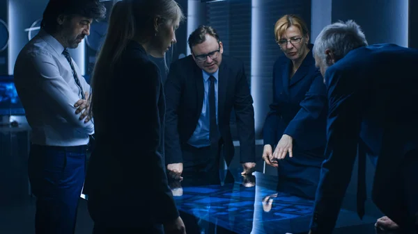 Equipo Diverso de Agentes Gubernamentales de Inteligencia Parados Alrededor de la Tabla de Pantalla Táctil Digital y el Sospechoso de Seguimiento, Oficial Superior hace Gestos Interactivos. Gran sala de vigilancia oscura. — Foto de Stock