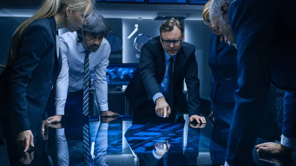 Diverse Team of Government Intelligence Agents standing Around Digital Touch Screen Table and Tracking Subject. Velká tmavá kontrolní místnost plná počítačových obrazovek. — Stock fotografie