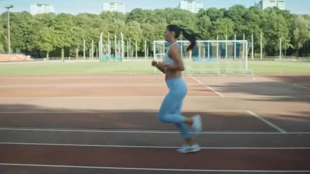 美丽的健身女人在浅蓝色运动上衣和在体育场慢跑。在一个温暖的夏日午后，她跑得很快。运动员做她的例行运动练习。跟踪拍摄. — 图库视频影像