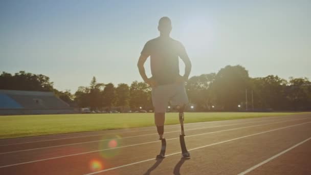 Спортсмен-инвалид с протезными бегущими лезвиями позирует во время тренировки на открытом стадионе в солнечный день. Ампутированный бегун стоит на треке. Спортивный след . — стоковое видео
