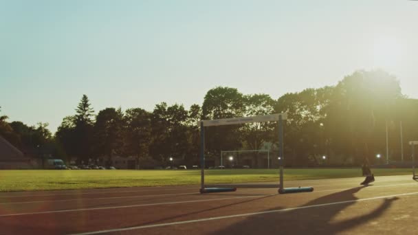 Атлетичний гравець в сірій куртці і короткометражці Хердлінг на стадіоні. Він перестрибує через бар'єри під час теплішого літнього дня. Спортсмен займається спортом на природі. — стокове відео