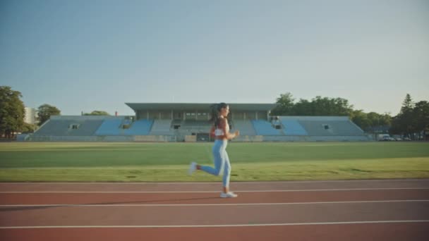 美丽的健身女人在浅蓝色运动上衣和在体育场慢跑。在一个温暖的夏日午后，她跑得很快。运动员做她的例行运动练习。跟踪拍摄. — 图库视频影像