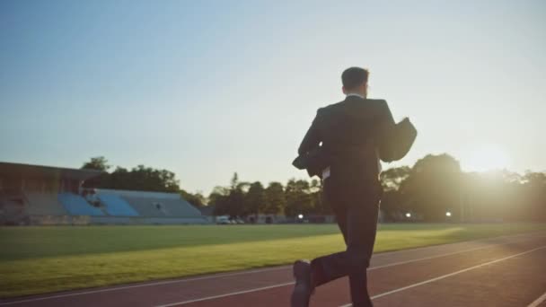 Jonge serieuze zakenman in een pak die in een buiten stadion loopt. Hij draagt een bril en houdt een mobiele telefoon. Kantoormedewerker die doelen achtervolgt. Management satire. Van achteren geschoten. — Stockvideo