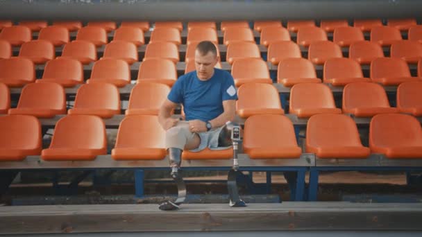 Sportowy Niepełnosprawny Fit Man z protetycznymi ostrzami do biegania przygotowuje się do treningu na stadionie w słoneczne popołudnie. Amputee Runner siedzi i utrwala nogi do biegu. — Wideo stockowe