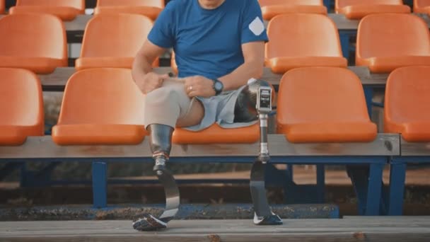 Athletic Disabled Fit Man with Prosthetic Running Blades se está preparando para un entrenamiento en un estadio al aire libre en una tarde soleada. Amputado corredor está sentado y fija sus piernas para una carrera . — Vídeo de stock