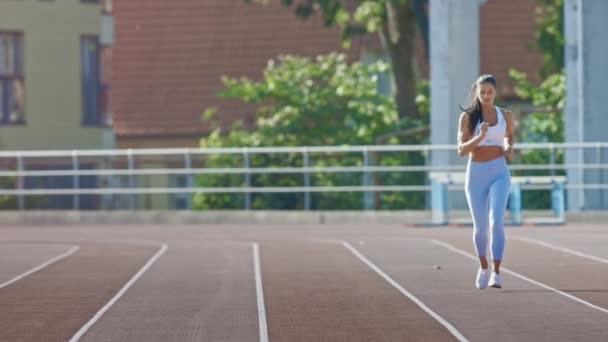 美丽的健身女人在浅蓝色运动上衣和在体育场慢跑。她在一个温暖的夏日午后跑步。运动员在跑道上进行日常体育锻炼。慢动作. — 图库视频影像