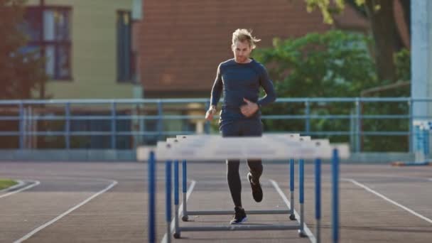 Athletic Fit Man in Grey Shirt and Shorts Hurdling in the Stadium (en inglés). Está saltando sobre barreras en una cálida tarde de verano. Atleta haciendo su práctica deportiva rutinaria. Tiro de seguimiento en cámara lenta . — Vídeo de stock