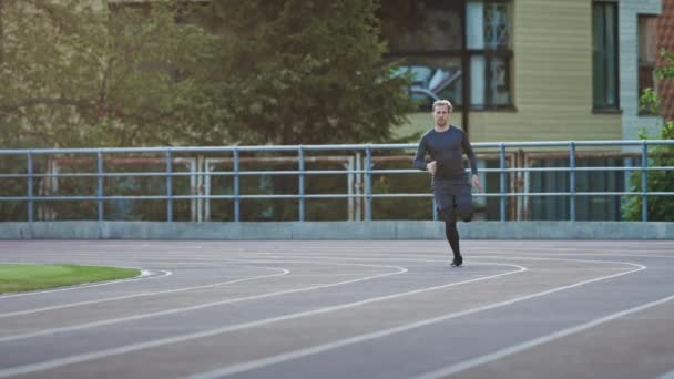 경기장에서 회색 셔츠와 반바지 조깅에 미소 운동 적합 남자. 그는 따뜻한 여름 오후에 빨리 달리고 있습니다. 그의 일상적인 스포츠 연습을 하 고 선수. 슬로우 모션 샷. — 비디오