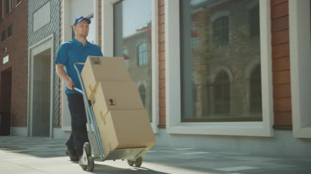 Happy Young Delivery Man Pushes kézi targonca tele kartondobozok és csomagok a szállítás. Professzionális futár hatékonyan és gyorsan dolgozik. A háttérben Stílusos modern városi terület — Stock videók